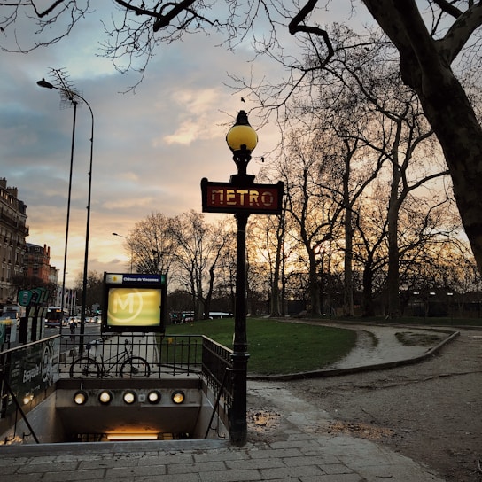 A Paris metro entrance is seen with a traditional art nouveau style sign, under a cloudy sky during sunset. Bare trees line the walkway, and a bicycle is parked near the entrance. The warm light from the streetlamps creates a serene and classic atmosphere.