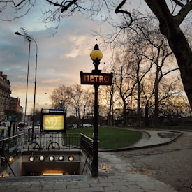 A Paris metro entrance is seen with a traditional art nouveau style sign, under a cloudy sky during sunset. Bare trees line the walkway, and a bicycle is parked near the entrance. The warm light from the streetlamps creates a serene and classic atmosphere.