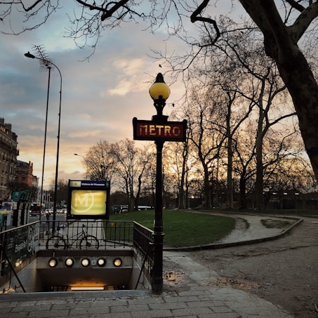 A Paris metro entrance is seen with a traditional art nouveau style sign, under a cloudy sky during sunset. Bare trees line the walkway, and a bicycle is parked near the entrance. The warm light from the streetlamps creates a serene and classic atmosphere.