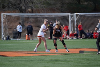 Two athletes are positioned on a lacrosse field, one wearing a white uniform with orange accents and the other wearing a black uniform with red details. They are holding lacrosse sticks, facing each other, ready to engage in play. A referee is nearby, and spectators are visible in the background, sitting or standing near the goalposts.