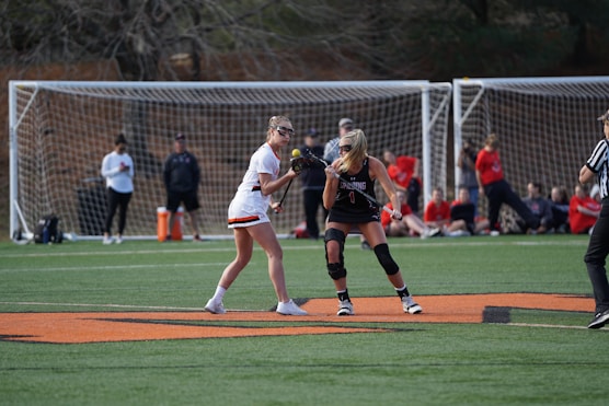 Two athletes are positioned on a lacrosse field, one wearing a white uniform with orange accents and the other wearing a black uniform with red details. They are holding lacrosse sticks, facing each other, ready to engage in play. A referee is nearby, and spectators are visible in the background, sitting or standing near the goalposts.