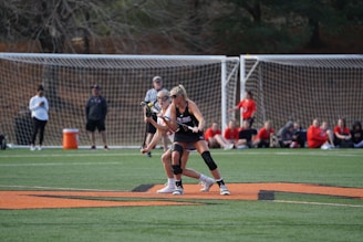 Two female athletes on a field are playing lacrosse, with one holding a lacrosse stick as they engage in the game. They are surrounded by a netted goal and a group of spectators and team members dressed in red sitting on the sidelines. The green field and an orange boundary line are also visible.