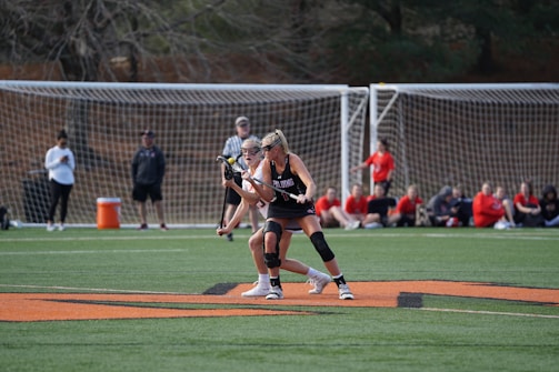 Two female athletes on a field are playing lacrosse, with one holding a lacrosse stick as they engage in the game. They are surrounded by a netted goal and a group of spectators and team members dressed in red sitting on the sidelines. The green field and an orange boundary line are also visible.