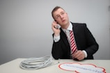 A person in a suit and striped tie is seated at a table, holding a phone to their ear and looking upward, appearing thoughtful or concerned. The table holds a coiled white cable and a cutout paper with red borders and text.