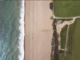An aerial view displaying a sandy beach alongside a body of water with visible white waves. Adjacent to the beach is a grassy area with a small parking lot containing several vehicles. A modest building is situated near the parking lot. A narrow pathway or pier extends slightly into the water.