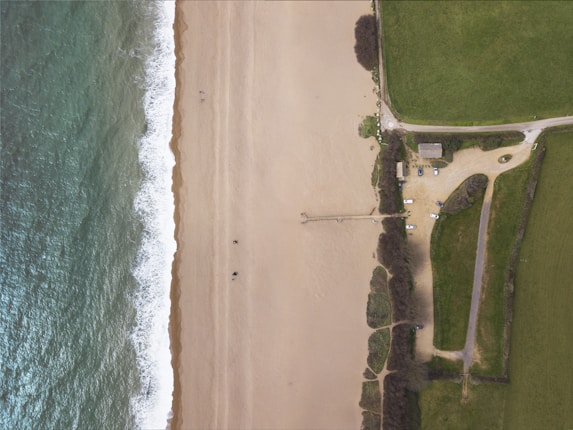 An aerial view displaying a sandy beach alongside a body of water with visible white waves. Adjacent to the beach is a grassy area with a small parking lot containing several vehicles. A modest building is situated near the parking lot. A narrow pathway or pier extends slightly into the water.