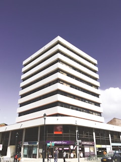 A modern, multi-story office building with white and glass exteriors. The structure has a sleek, geometric design with several uniform layers. At the base, there are storefronts with signage, and a few pedestrians are visible on the sidewalk. The sky is clear and slightly purple, creating an interesting backdrop.