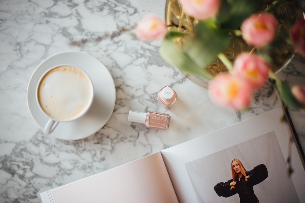 coffee on cup with saucer beside nail polish bottles, book, and flowers