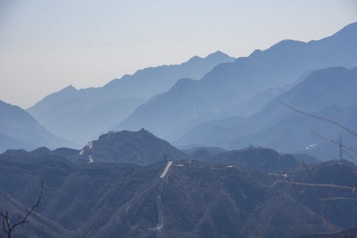 A series of majestic mountains extend into the distance, veiled in a soft, bluish haze. The iconic Great Wall of China winds through the rugged terrain, creating a sense of historical depth and grandeur.