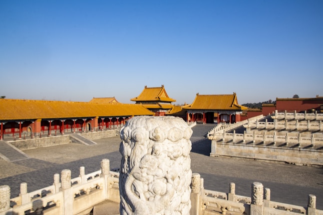 A majestic ancient Chinese palace complex featuring traditional architecture with ornate yellow-tiled roofs and red walls. In the foreground, an intricately carved stone pillar stands among the elevated walkways and steps, surrounded by white stone railings.