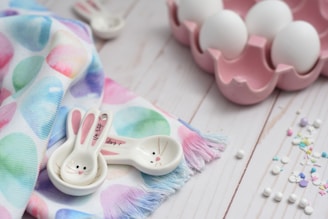 Colorful fabric with pastel egg patterns lies beside ceramic measuring spoons shaped like bunny faces. Nearby, a pink egg holder contains white eggs, and small sprinkles are scattered on a light wooden surface.