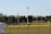 A sports field with a group of people engaging in athletic activities in the background. There is sports equipment like a soccer goal and a scoreboard visible. A chain-link fence runs across the foreground, and a statue or sculpture resembling an eagle is also seen. Trees and tall floodlights are positioned behind the field.