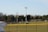 A sports field with a group of people engaging in athletic activities in the background. There is sports equipment like a soccer goal and a scoreboard visible. A chain-link fence runs across the foreground, and a statue or sculpture resembling an eagle is also seen. Trees and tall floodlights are positioned behind the field.