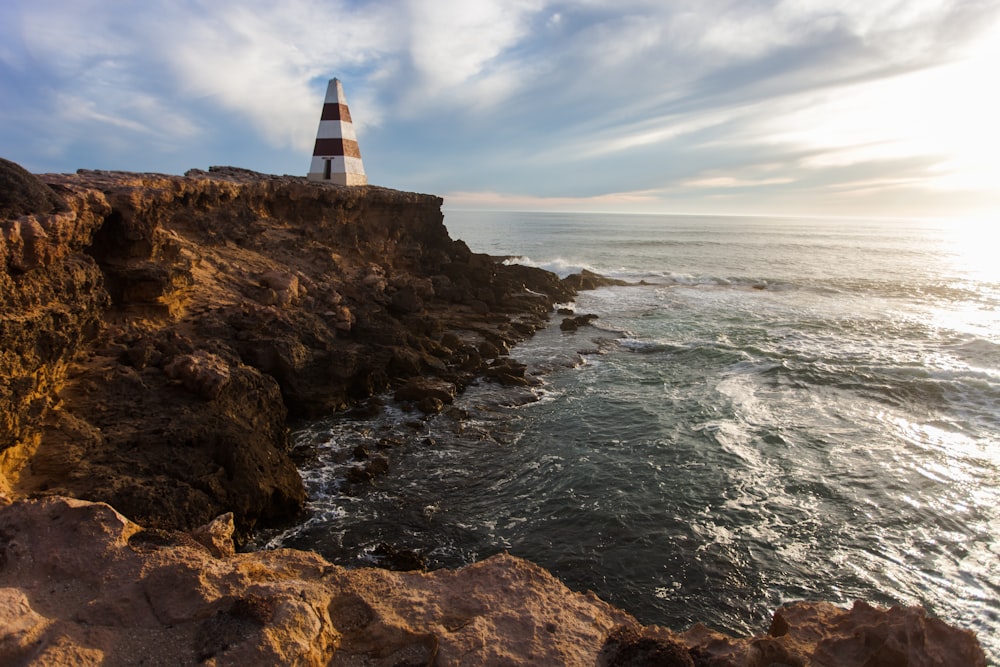 brown and white lighthouse on rocky cliff facing ocean