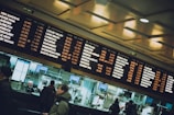 A busy train station with large display boards listing train destinations, departure times, and tracks. Several people are visible in the foreground, interacting with ticket counters that have numbered windows. The lighting is warm with reflections on the glass enclosures.