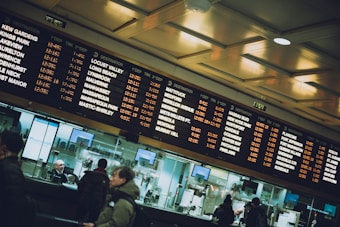 A busy train station with large display boards listing train destinations, departure times, and tracks. Several people are visible in the foreground, interacting with ticket counters that have numbered windows. The lighting is warm with reflections on the glass enclosures.