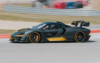 A sleek, high-performance sports car speeding on a racetrack. The car features a black and gold color scheme, with aerodynamic elements like a large rear wing and low-profile tires. The background shows blurred race barriers and grassy areas, indicating motion.