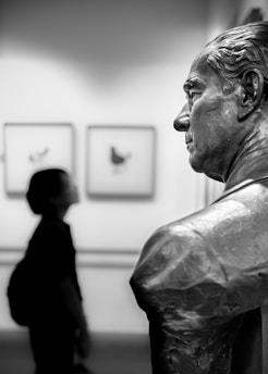 A black and white photograph depicting a close-up of a bronze statue of a man's upper body and face, with detailed features. In the background, a person with short hair is observing the artwork, holding a bag on their shoulder. Two framed artworks featuring abstract shapes are visible on the wall.