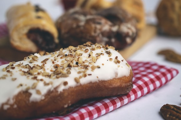 A pastry with a white icing is topped with crushed nuts, placed on a red and white checkered cloth. Other baked goods are blurred in the background, creating a cozy and inviting setting.