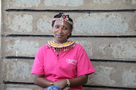 A person wearing a vibrant traditional beaded necklace and headpiece sits against a stone wall. They are wearing a pink polo shirt with a logo that reads 'Dignit'. The design of the necklace and headpiece is ornate and colorful, contrasting with the textured stone background.
