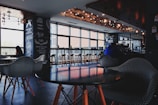 A cozy cafe interior with modern design elements, featuring a series of wooden tables and chairs arranged near a large window. Several patrons are seated, enjoying their drinks, with pendant lighting providing a warm ambiance. Decor elements include chalkboard-style menu boards on the wall.