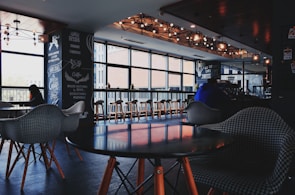 A cozy cafe interior with modern design elements, featuring a series of wooden tables and chairs arranged near a large window. Several patrons are seated, enjoying their drinks, with pendant lighting providing a warm ambiance. Decor elements include chalkboard-style menu boards on the wall.
