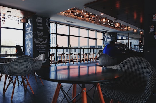 A cozy cafe interior with modern design elements, featuring a series of wooden tables and chairs arranged near a large window. Several patrons are seated, enjoying their drinks, with pendant lighting providing a warm ambiance. Decor elements include chalkboard-style menu boards on the wall.