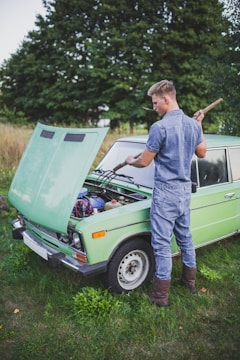A person dressed in denim is working on the engine of an old green car with the hood open, set against a backdrop of trees and grass.