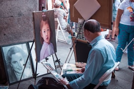 An artist is seated and working on a painting of a young girl. The painting is displayed on an easel, and there is another portrait of a woman leaning against the wall. Several people are standing and sitting in the background, engaged in different activities.
