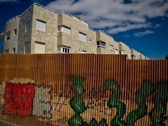 A residential building with a stone exterior stands behind a corrugated metal fence adorned with graffiti. The sky above is partly cloudy, enhancing the contrast with the building's light facade.