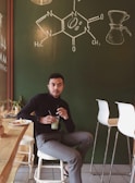 A person sits in a cozy cafe environment with a green chalkboard wall displaying a chemical structure and a coffee brewing diagram. The individual is holding a glass with a green beverage, with a plate of snacks nearby. The setting includes modern bar stools and a light wooden countertop, contributing to a relaxed and inviting atmosphere.