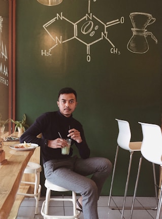 A person sits in a cozy cafe environment with a green chalkboard wall displaying a chemical structure and a coffee brewing diagram. The individual is holding a glass with a green beverage, with a plate of snacks nearby. The setting includes modern bar stools and a light wooden countertop, contributing to a relaxed and inviting atmosphere.