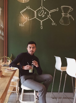 A person sits in a cozy cafe environment with a green chalkboard wall displaying a chemical structure and a coffee brewing diagram. The individual is holding a glass with a green beverage, with a plate of snacks nearby. The setting includes modern bar stools and a light wooden countertop, contributing to a relaxed and inviting atmosphere.
