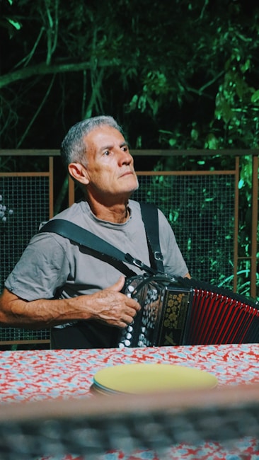 An elderly man with gray hair sits at a brightly colored table holding an accordion. He appears to be deep in thought, looking slightly upwards. The setting is outdoors, with a backdrop of dense green foliage and a wooden fence.