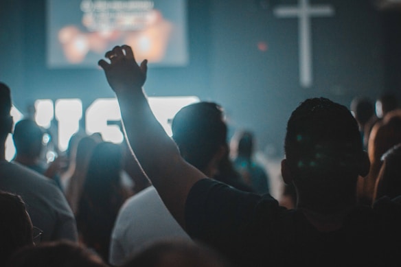 A group of people are gathered in a dimly lit room, with one individual raising a hand towards the front. A large illuminated cross is visible on the wall, suggesting a religious setting. The atmosphere appears contemplative or worshipful, with most figures facing away.