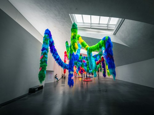 A spacious gallery with high ceilings displays a large, hanging art installation made of vibrant, multicolored fabric. The fabric pieces are arranged to resemble flowing strands, resembling a surreal, colorful forest. A few people are admiring the installation, adding a sense of scale to the artwork.