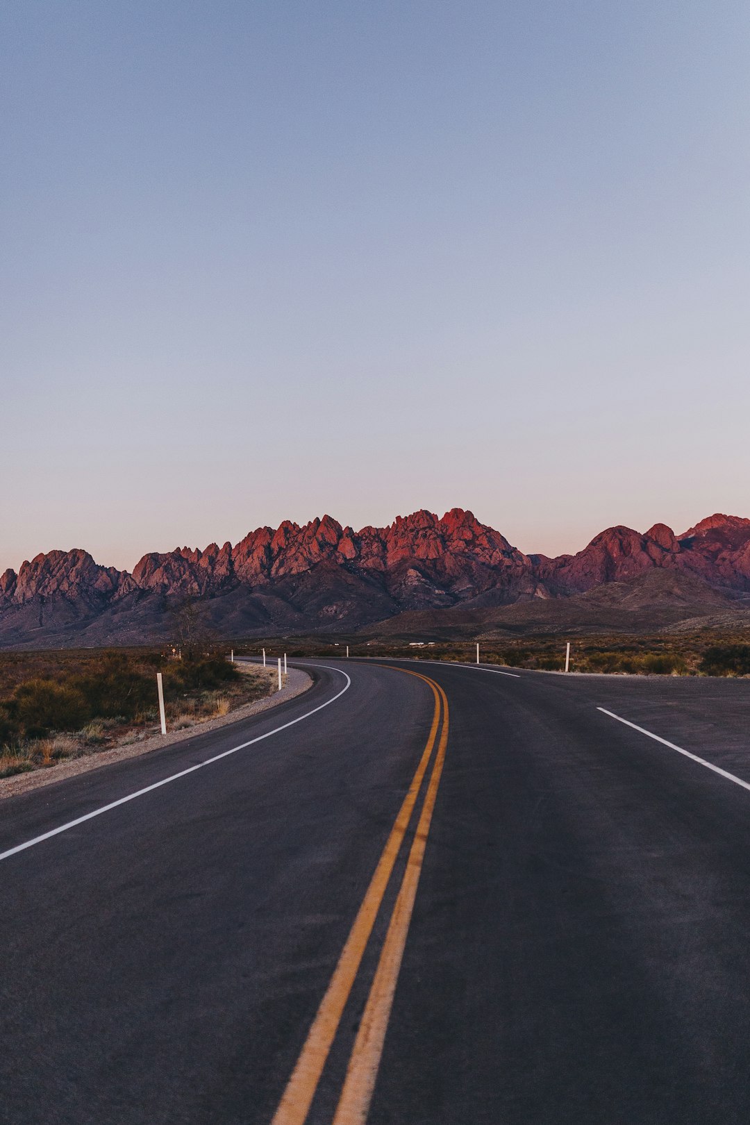 The image shows the Sandia mountains in New Mexico at sunset when the mountains and sky turn a  beautiful pink, purple and blue. A road, free from any traffic is also pictured, The road goes towards the mountain. 