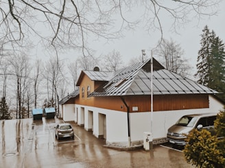 A building with a triangular roof featuring solar panels is situated in a parking area. Two parked cars are visible next to it. Leafless trees surround the area, and the ground appears wet, suggesting recent rain.