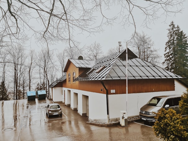 A building with a triangular roof featuring solar panels is situated in a parking area. Two parked cars are visible next to it. Leafless trees surround the area, and the ground appears wet, suggesting recent rain.