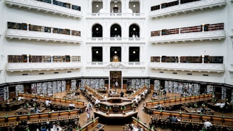 A grand, multi-level library with ornate architecture and balustrades. The space is filled with rows of long wooden tables and green lamps. Bookshelves line the walls and are filled with numerous books. The setting is busy with people reading, studying, and working.