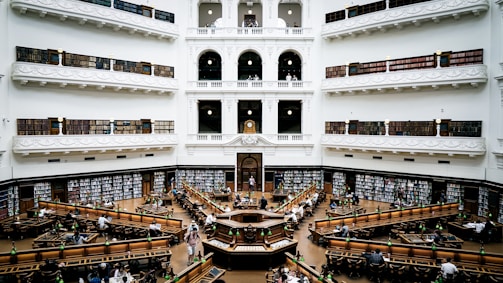 A grand, multi-level library with ornate architecture and balustrades. The space is filled with rows of long wooden tables and green lamps. Bookshelves line the walls and are filled with numerous books. The setting is busy with people reading, studying, and working.