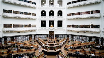 A grand, multi-level library with ornate architecture and balustrades. The space is filled with rows of long wooden tables and green lamps. Bookshelves line the walls and are filled with numerous books. The setting is busy with people reading, studying, and working.