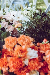 Various flowers including orange marigolds, white and pink roses, and white daisies are arranged densely with green foliage surrounding them. In the background, there are light blue folding chairs, likely set up for an event outdoors.