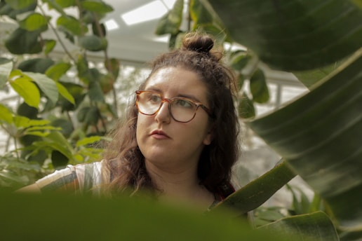 A person with long, wavy hair and glasses is surrounded by lush green foliage, creating a serene and contemplative atmosphere.