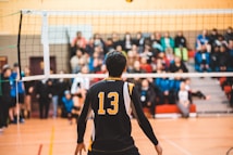 A volleyball player is standing on an indoor court, wearing a black and yellow sports jersey with the number 13. The player faces the net, focusing on the game. In the background, spectators are seated on bleachers, watching the match with interest.