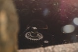 A close-up of a rusty, metallic gear situated in a dark, industrial setting. The gear is central in focus, with a surrounding structure that appears weathered and aged. Soft light filters through, creating a contrast between the shadows and highlights, adding an element of mystery to the scene.