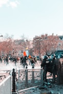 A scene of civil unrest featuring a group of riot police in protective gear positioned behind barricades facing protesters. The protesters, some carrying flags and signs, are moving through a street littered with debris. Nearby, leafless trees and traditional European architecture suggest an urban setting. The atmosphere is tense and chaotic.
