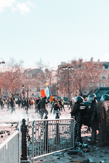 A scene of civil unrest featuring a group of riot police in protective gear positioned behind barricades facing protesters. The protesters, some carrying flags and signs, are moving through a street littered with debris. Nearby, leafless trees and traditional European architecture suggest an urban setting. The atmosphere is tense and chaotic.