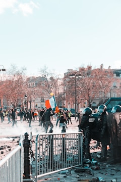 A scene of civil unrest featuring a group of riot police in protective gear positioned behind barricades facing protesters. The protesters, some carrying flags and signs, are moving through a street littered with debris. Nearby, leafless trees and traditional European architecture suggest an urban setting. The atmosphere is tense and chaotic.