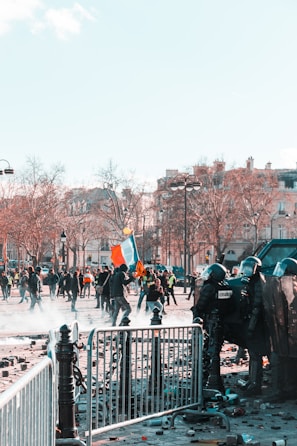 A scene of civil unrest featuring a group of riot police in protective gear positioned behind barricades facing protesters. The protesters, some carrying flags and signs, are moving through a street littered with debris. Nearby, leafless trees and traditional European architecture suggest an urban setting. The atmosphere is tense and chaotic.